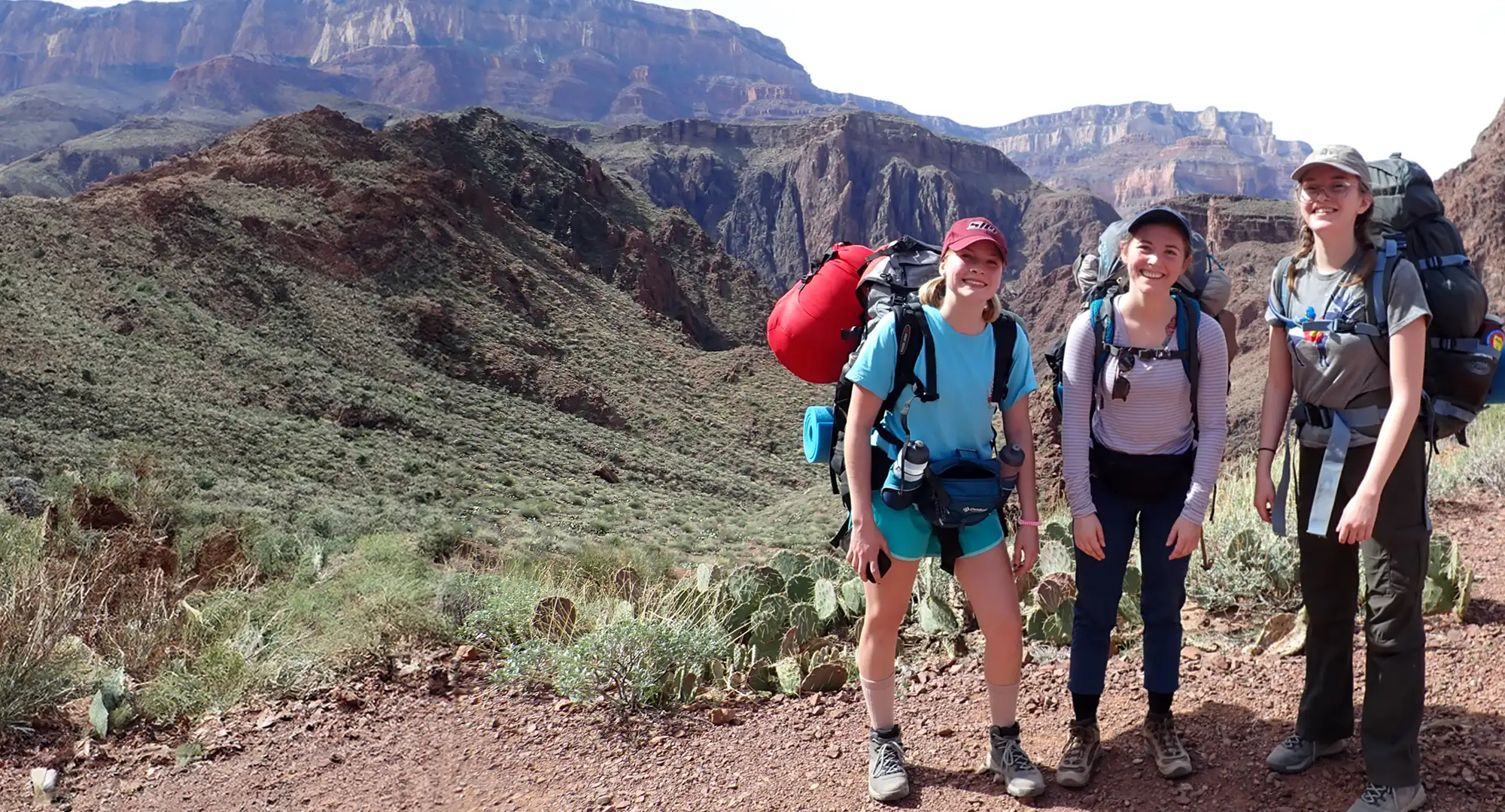 Three hikers with large backpacks stand on a dirt trail, smiling at the camera. Behind them is a rugged desert landscape with cliffs, rocky hills, and sparse vegetation under a clear sky.